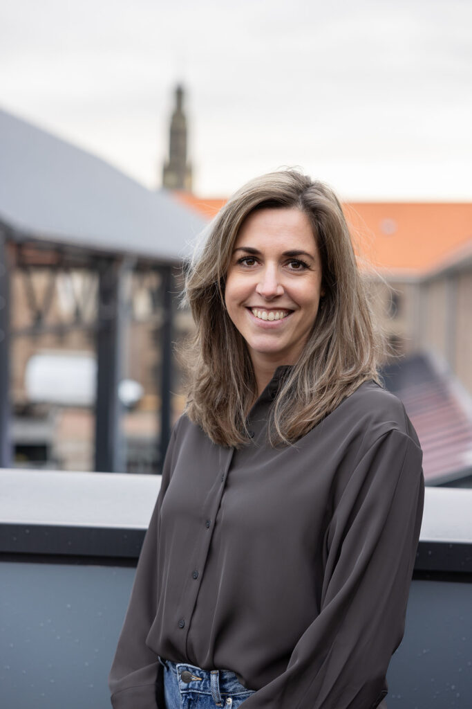 Portretfoto van Renée Wolters – Seegers op een dakterras bij vergaderlocatie Onder de Pannen in Echt, met een industrieel gebouw en een kerktoren op de achtergrond.