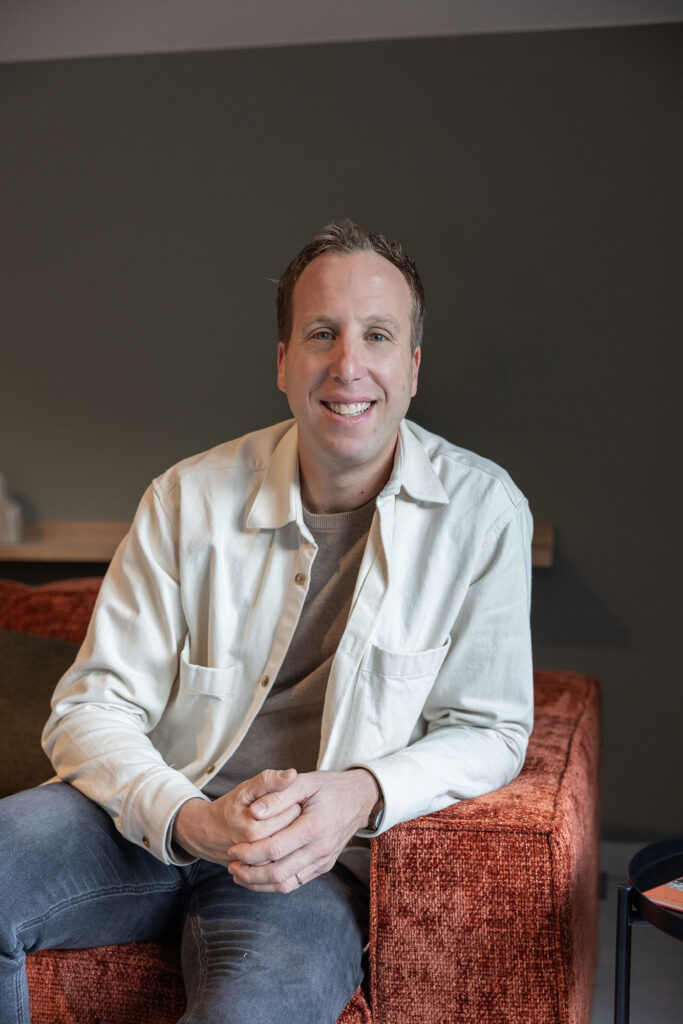 Portretfoto van Joop Wolter in een moderne, sfeervolle vergaderruimte bij Onder de Pannen in Echt. Hij zit ontspannen op een roestkleurige fauteuil, met een groene muur en houten accenten op de achtergrond.